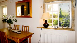 A dining area with a wooden table, chairs, a vase of flowers, a lamp, and a window with a view of greenery.