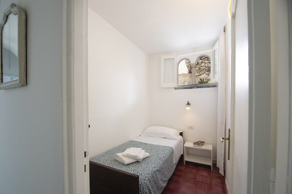Una habitación pequeña y limpia con una cama individual, una mesita de noche, un espejo y un estante pequeño con un objeto decorativo.