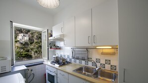 A compact kitchen with white cabinets, a stainless steel sink, and a window overlooking a hillside. - Blessed villa directly on the sea. (Praiano)