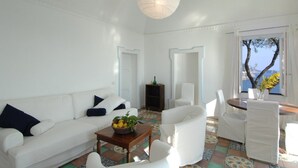 A bright living room with a white sofa, a wooden coffee table with a fruit bowl, white armchairs, and a view of the sea through a window. - Blessed villa directly on the sea. (Praiano)