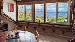 A dining area with a table and chairs, large windows with a view of the ocean and hills, and a wooden ceiling.