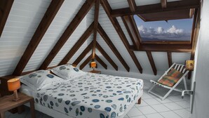 A bedroom with a sloped ceiling, a bed with patterned bedding, a bedside table with a lamp, a window with a view of the sky and clouds, and a small chair with a cushion.
