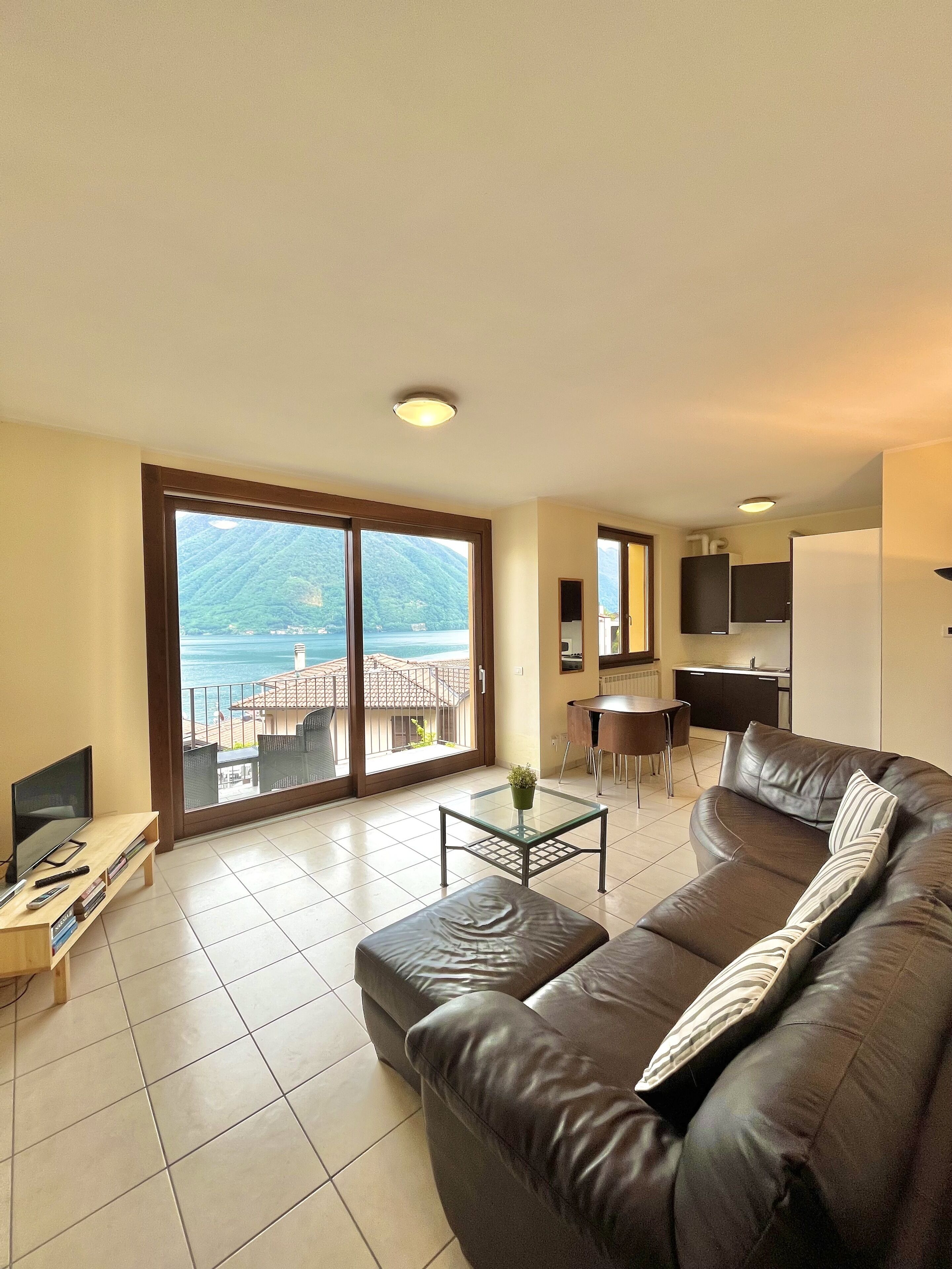 A living room with a large sofa, a coffee table, and a TV. There is a balcony with a view of the sea and mountains.
