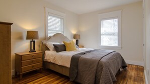 Une chambre à coucher comprenant un lit, deux tables de chevet, une commode et deux fenêtres équipées de stores.