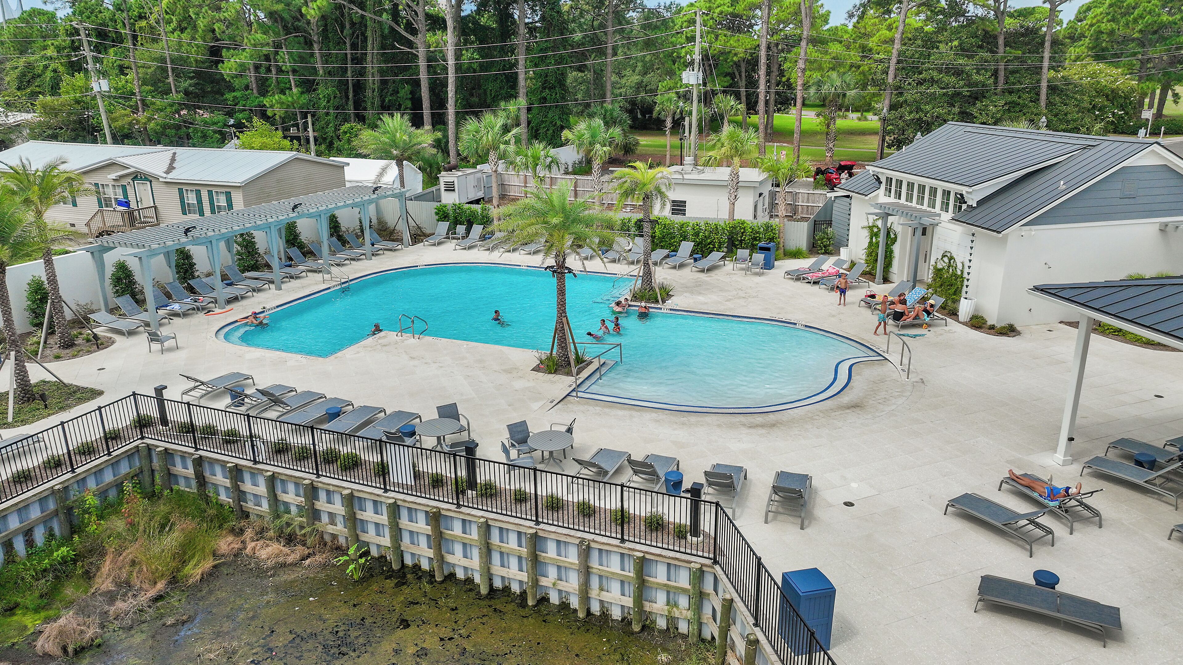 Une piscine entourée de bâtiments et d’une zone délimitée par une clôture, avec des chaises longues et des palmiers.