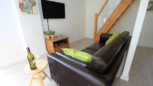 Una sala moderna con un sofá de cuero, una mesita de madera, un televisor montado en la pared y una escalera con barandilla de vidrio.