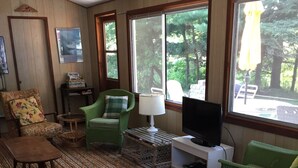 A cozy living room with green wicker furniture, a television, and large windows offering a view of trees. - Cottage For Rent-1000 Islands (Gananoque)