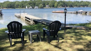A lakeside deck with two Adirondack chairs and a small table, overlooking a calm lake with boats and houses in the background. - Cottage For Rent-1000 Islands (Gananoque)