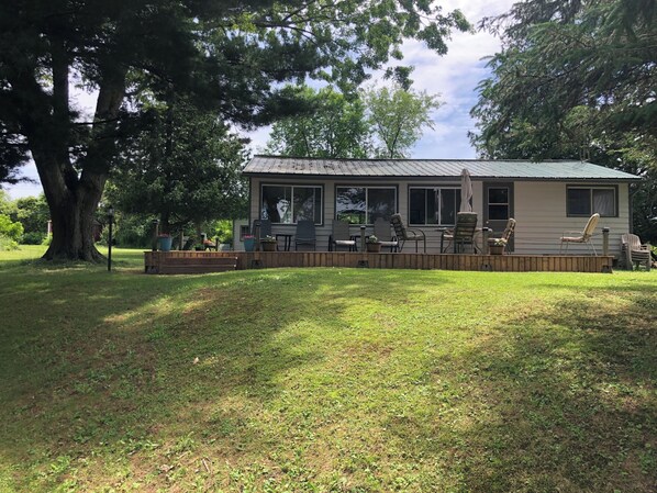 A single-story house with a covered patio area, a large tree in the foreground, and a clear sky. - Cottage For Rent-1000 Islands (Gananoque)