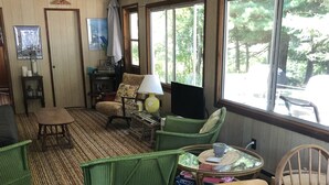 A cozy living room with a wooden table, green chairs, a patterned rug, and a window with a view of trees. - Cottage For Rent-1000 Islands (Gananoque)