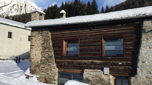 A rustic log cabin with stone walls and wooden windows, surrounded by snow-covered ground and a mountainous backdrop. - Rustico with style, modern and rustic, 2-4 P, in Val Poschiavo (Poschiavo)