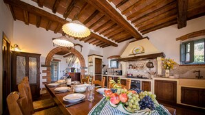 Un coin repas avec une longue table en bois dressée pour le repas, une cuisine en arrière-plan et un plafond rustique avec des poutres apparentes.