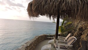Un salón junto a la playa con sillones blancos y una sombrilla de paja, con vistas al océano.