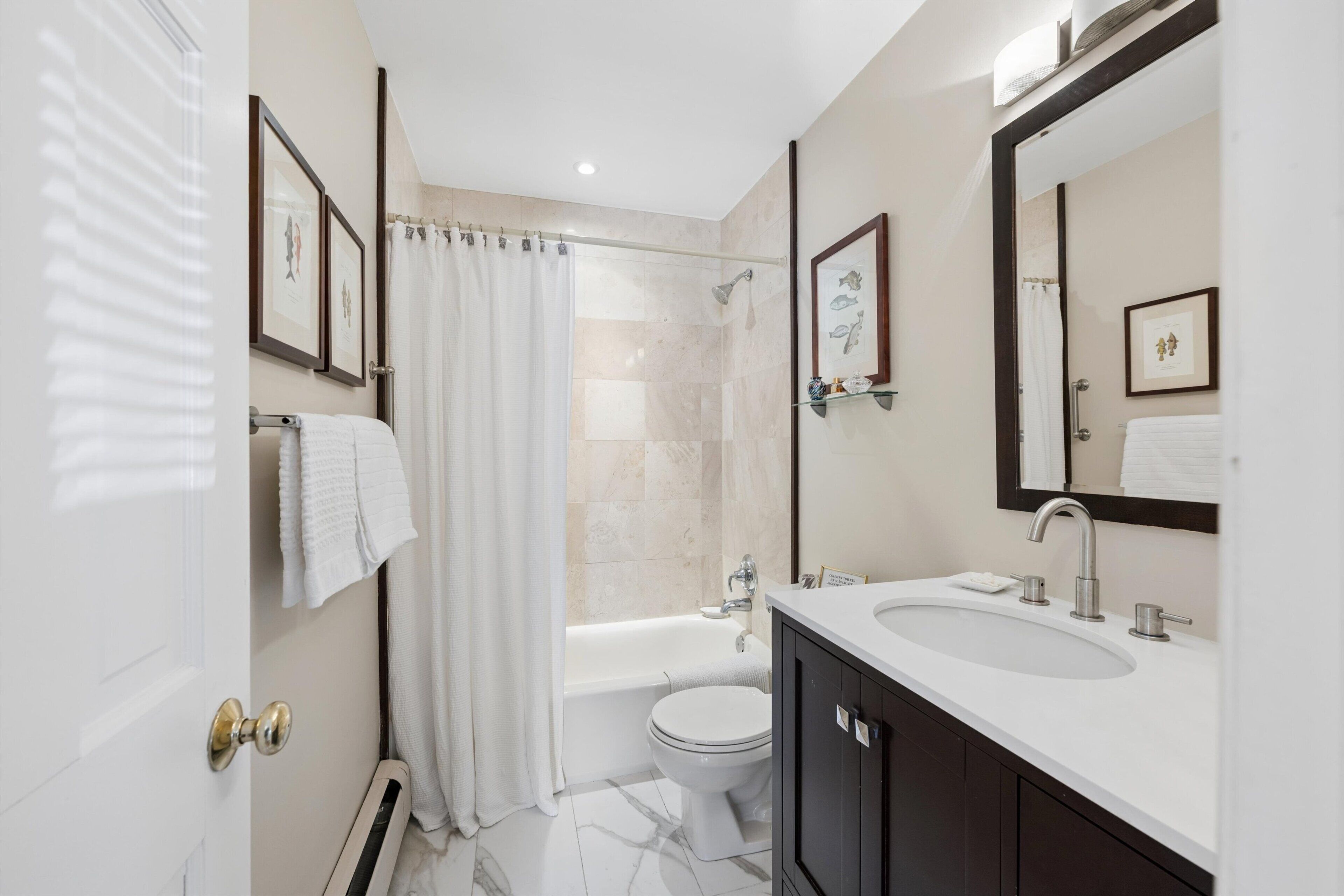 A bathroom with a bathtub, toilet, and sink, featuring a shower curtain, framed artwork, and a large mirror.