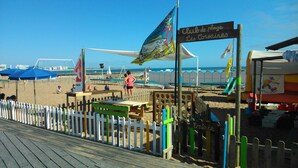 En træterrasse ved stranden med et hvidt stakittfence, et skilt der siger "Club de plage Les Corsaires", og et strandområde med parasoller og mennesker.