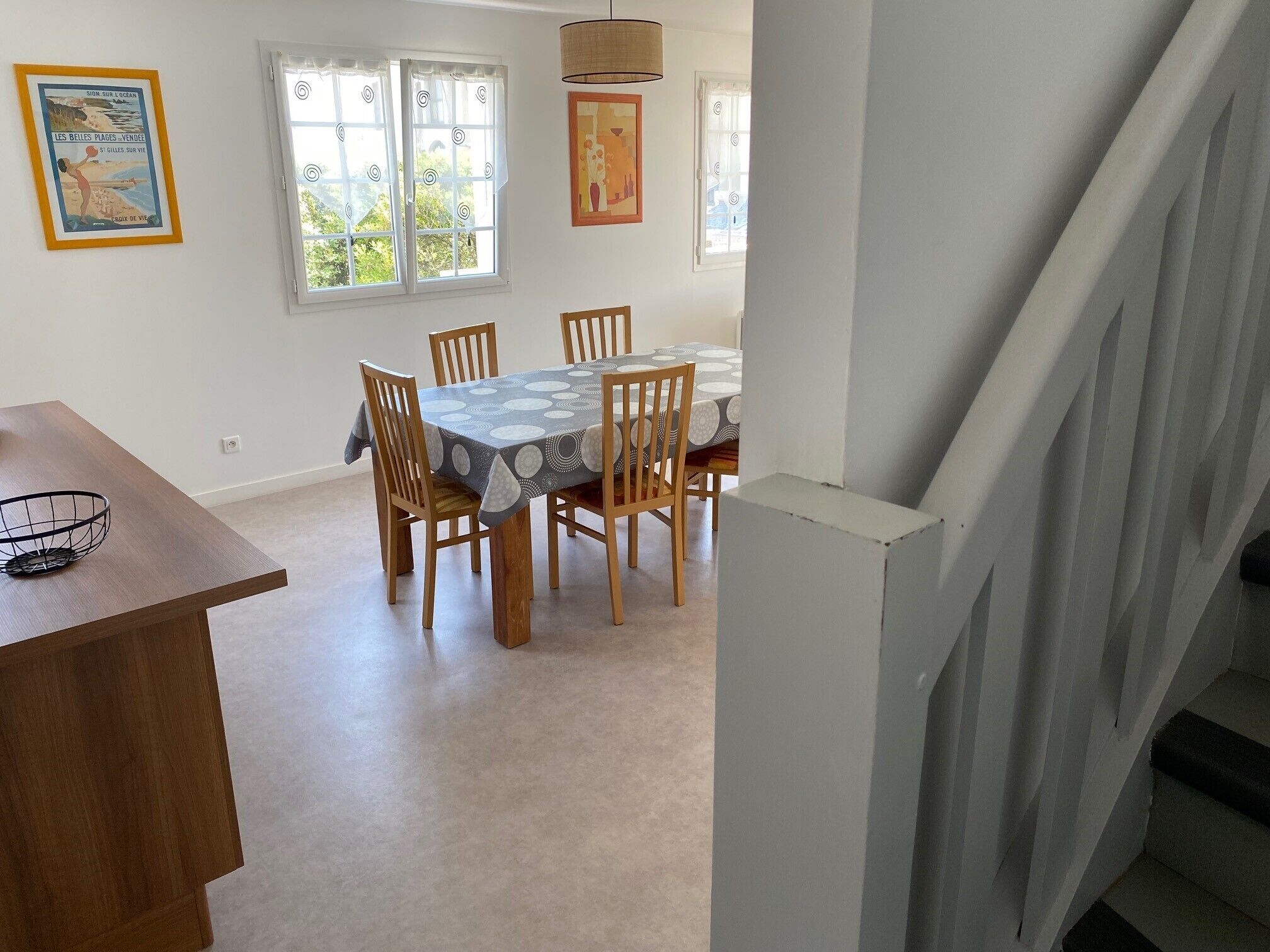 A dining area with a table set for six, wooden chairs, a patterned tablecloth, and a window with curtains.