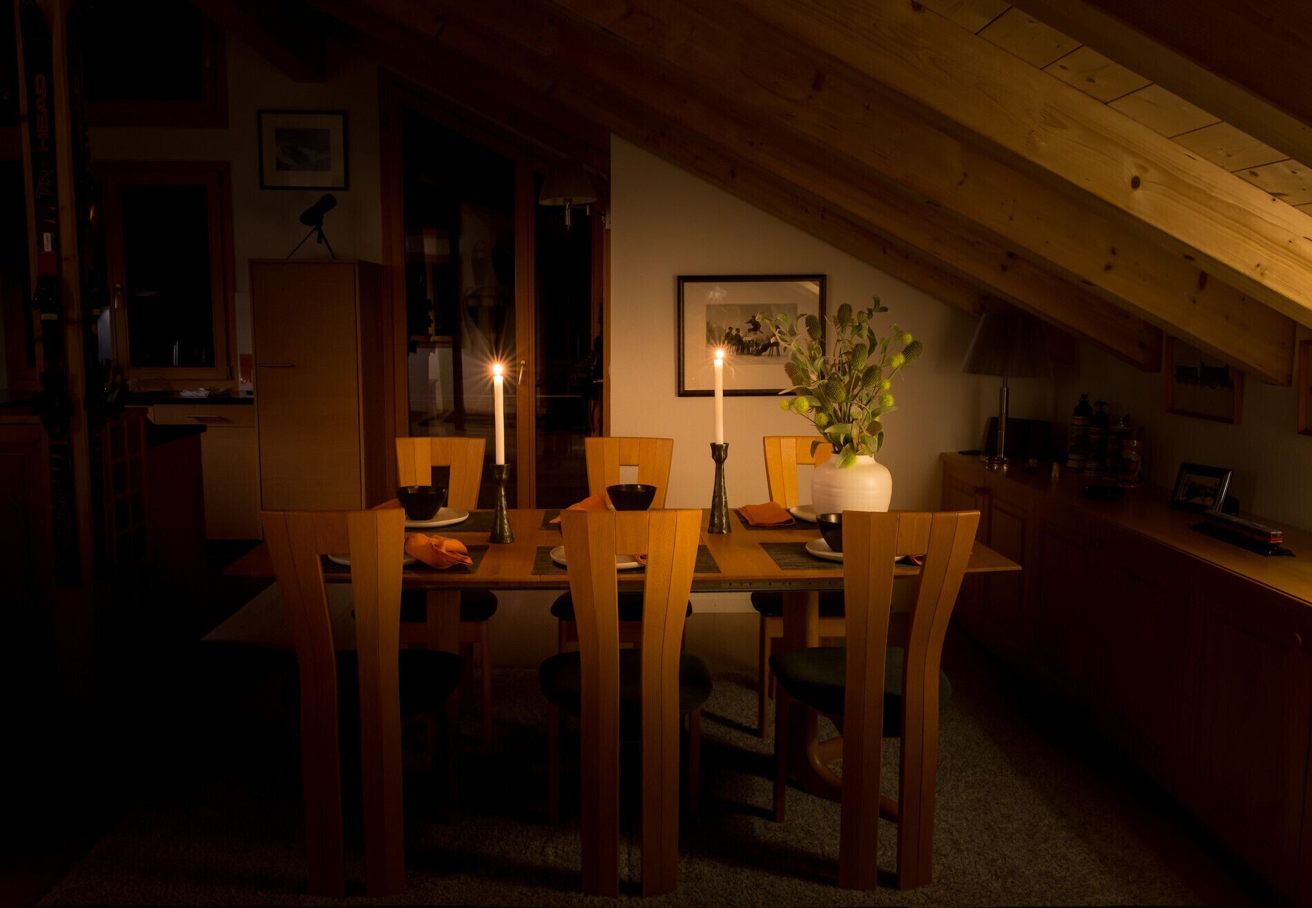 A dimly lit dining area with a table set for four, featuring a candle, a vase with green plants, and a framed picture on the wall.