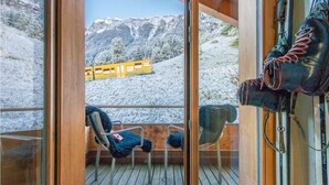 Una terraza de madera con sillas y un soporte para botas de esquí, con vistas a una montaña nevada y un tren amarillo.
