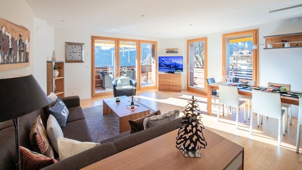 Ein geräumiges Wohnzimmer mit einer Couch, einem Couchtisch und einem Fernseher. Es gibt einen Essbereich mit Tisch und Stühlen. Das Zimmer hat große Fenster mit Bergblick.