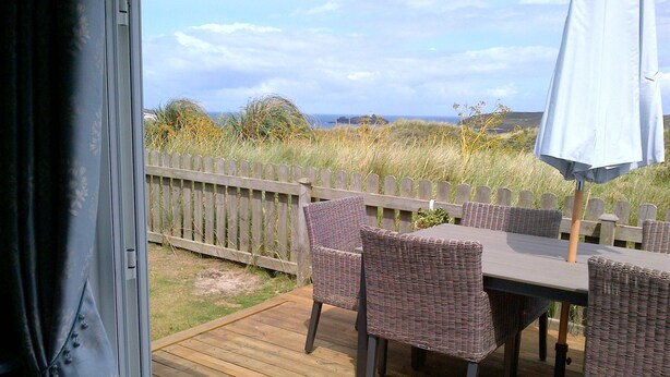 Een terras buiten met rieten meubels en een tafel, met uitzicht op een gazon met houten omheining en zee.