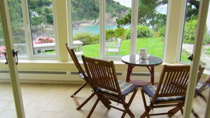 A room with large windows, two wooden chairs with cushions, a small round table with a pitcher and glass, and a view of a garden and water. - Detached Cottage Overlooking The Sea (Lee)
