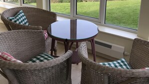 A room with wicker furniture, a glass table, and large windows offering a view of a green landscape and a body of water. - Detached Cottage Overlooking The Sea (Lee)