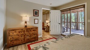A bedroom with a wooden dresser, a bed with a floral bedspread, a red and white rug, a ceiling fan, and a door leading to a balcony with a view of trees.