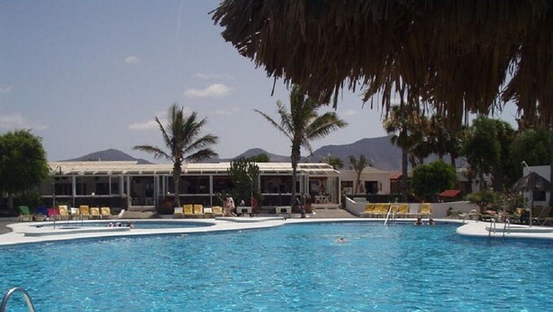 A swimming pool with lounge chairs and palm trees, surrounded by buildings and mountains in the background. - Villa 83 on the complex of Las Brisas  (Playa Blanca)