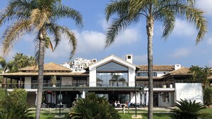 A two-story building with a flat roof, large windows, and a balcony overlooking a grassy area with palm trees.