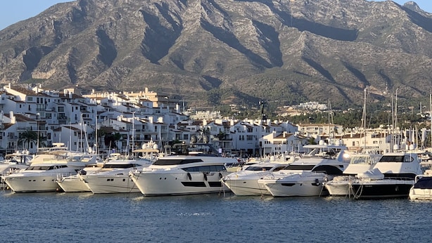 A marina with yachts docked in front of a mountainous backdrop.