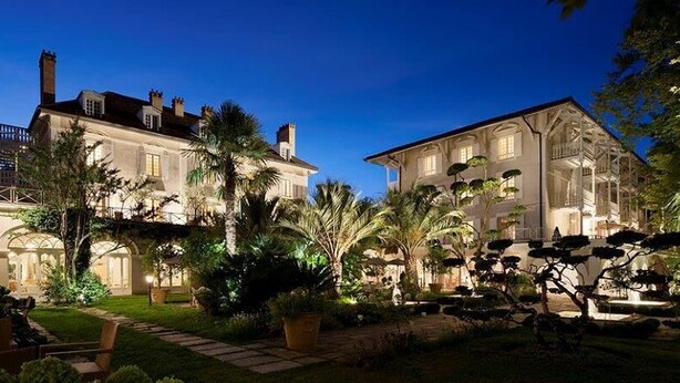 Un edificio grande e histórico de varios pisos, un jardín bien cuidado con palmeras y un camino empedrado que conduce a la entrada.