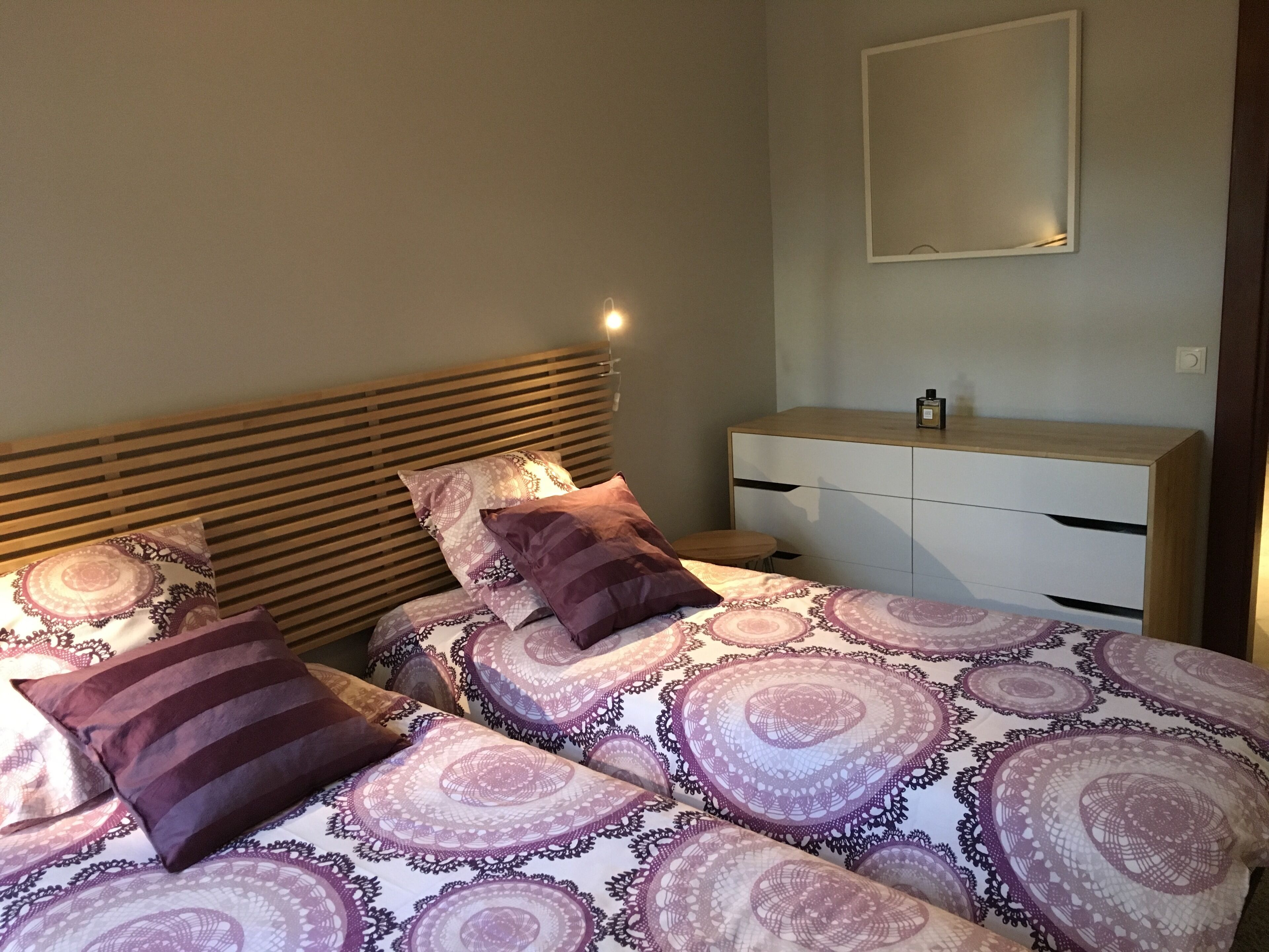 Une chambre avec deux lits, chacun agrémenté de draps et d’oreillers à motifs. On y trouve une table de chevet sur laquelle est posée une bouteille, ainsi qu’un petit tabouret. Un miroir est accroché au mur, au-dessus du lit.