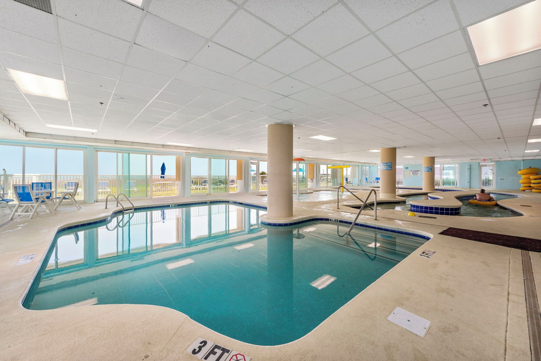 Ein Hallenbad mit klarem Wasser, einer Markierung für 90 Zentimeter Wassertiefe und Blick durch große Fenster nach draußen.