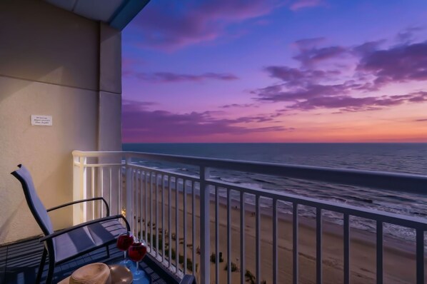 Uma varanda com vista para o oceano ao pôr do sol, uma cadeira e uma mesa com copos de vinho.