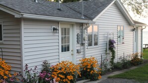 Una casa a un piano bianca con un portico, un piccolo giardino con fiori arancioni e rosa, e una cassetta delle lettere ben visibile.