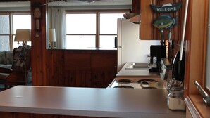 Cucina con mobili in legno, un piano di lavoro bianco, un fornello e un lavandino. Si nota una finestra con tende, una sveglia e un cartello di benvenuto con un pesce.