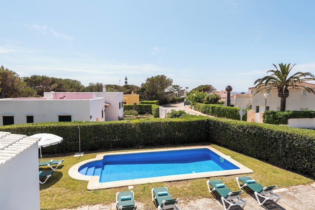 A swimming pool with lounge chairs, surrounded by neatly trimmed hedges and buildings in the background.