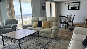 Un salon avec un canapé beige, un fauteuil gris, une table basse et un coin repas comprenant une table et des chaises.