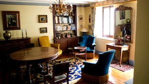 A traditional room with a wooden dining table, blue armchairs, a bookshelf, and a chandelier.