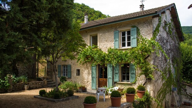 A stone house with green shutters, a gravel driveway, and potted plants.