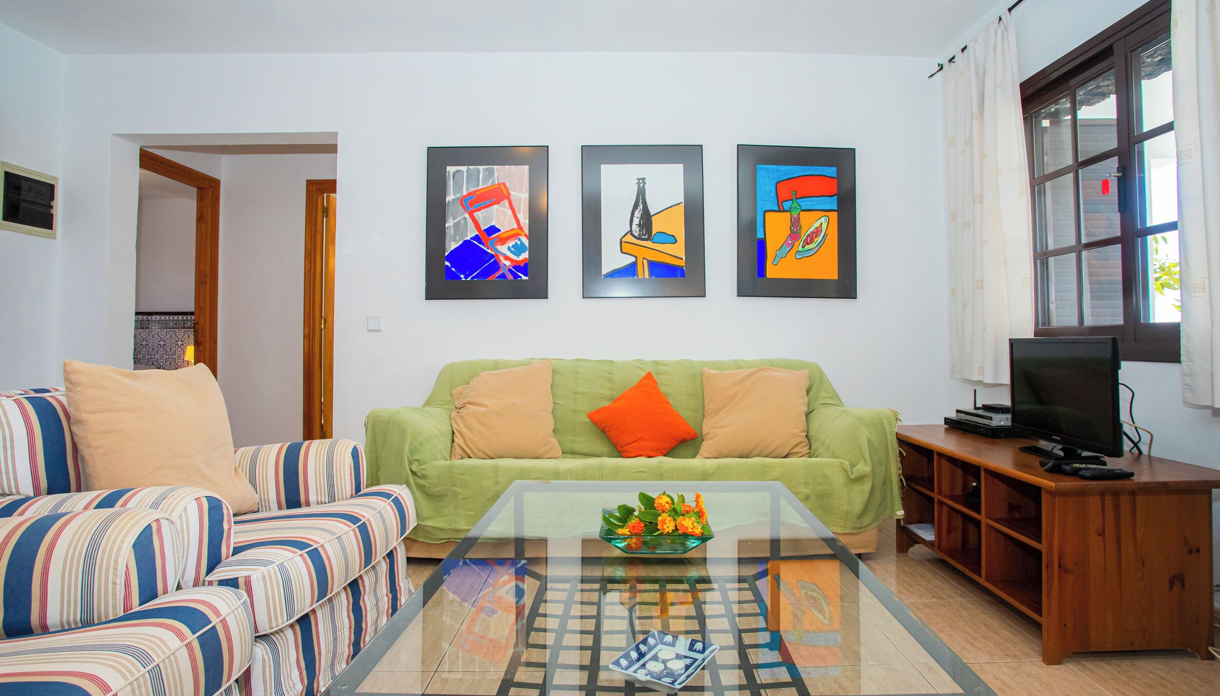 Ein Wohnzimmer mit einem gestreiften Sofa, einem grünen Sofa mit orangefarbenen Kissen, einem Glastisch mit Blumenvase und einem Fernseher auf einem Holzständer.
