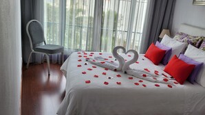 Una habitación de hotel con una cama decorada con pétalos de rosa y dos almohadas con forma de cisne, una silla gris y una ventana con cortinas translúcidas.