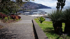 Un paseo marítimo junto a la costa, con un camino adoquinado, un puerto deportivo con barcos y montañas al fondo.