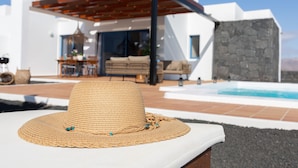 A modern house with a pool, a covered patio, and a lounge area with a hat on a chair.