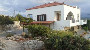 A modern white building with a red-tiled roof, stone pathway, and landscaped garden with rocks and shrubs. - Villa in a unique location with pool, beautiful gardens mountain and sea views. (Plaka)
