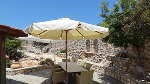 An outdoor patio with a table and chairs under a large umbrella, surrounded by stone walls and greenery. - Villa in a unique location with pool, beautiful gardens mountain and sea views. (Plaka)