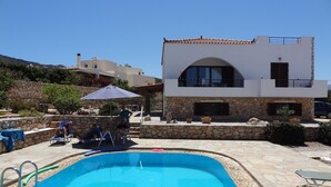 A swimming pool with a stone border, a stone wall, and a modern building with a white facade and a tiled roof. - Villa in a unique location with pool, beautiful gardens mountain and sea views. (Plaka)