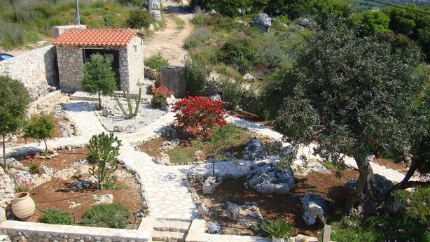 A stone pathway leading to a small stone building with a red-tiled roof, surrounded by a garden with various plants and a cactus. - Villa in a unique location with pool, beautiful gardens mountain and sea views. (Plaka)
