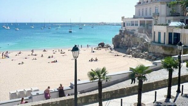 Une plage avec un bâtiment donnant sur la mer, agrémentée d’une promenade pavée et de palmiers.