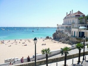 A beach with a building overlooking the sea, featuring a paved walkway and palm trees.